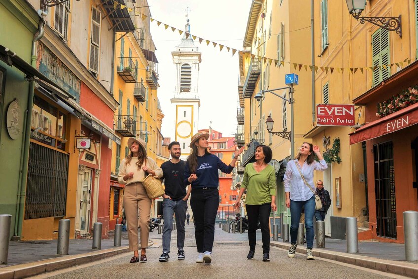 A group of people walking down a street lined with colorful buildings, a church spire in the background, and a sign for 'EVEN Prêt à Porter'. 2
