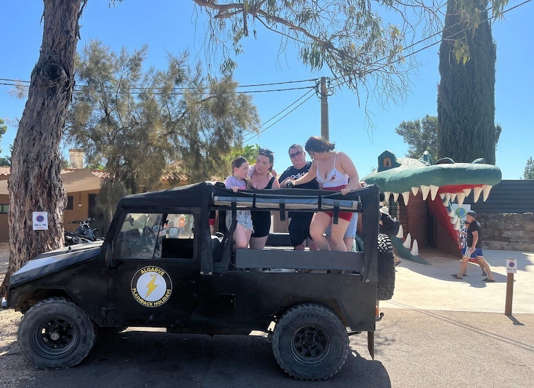 A black off-road vehicle with people sitting on top, parked near a large tree and a building. 2