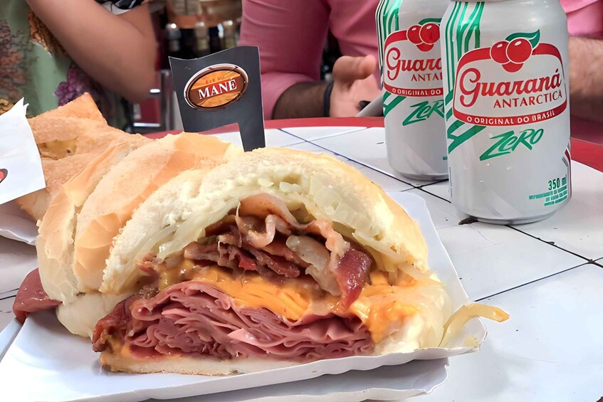 A sandwich with multiple layers of meat and cheese, a bottle of Guarana Antarctica Zero, and a Mané coffee cup. 5