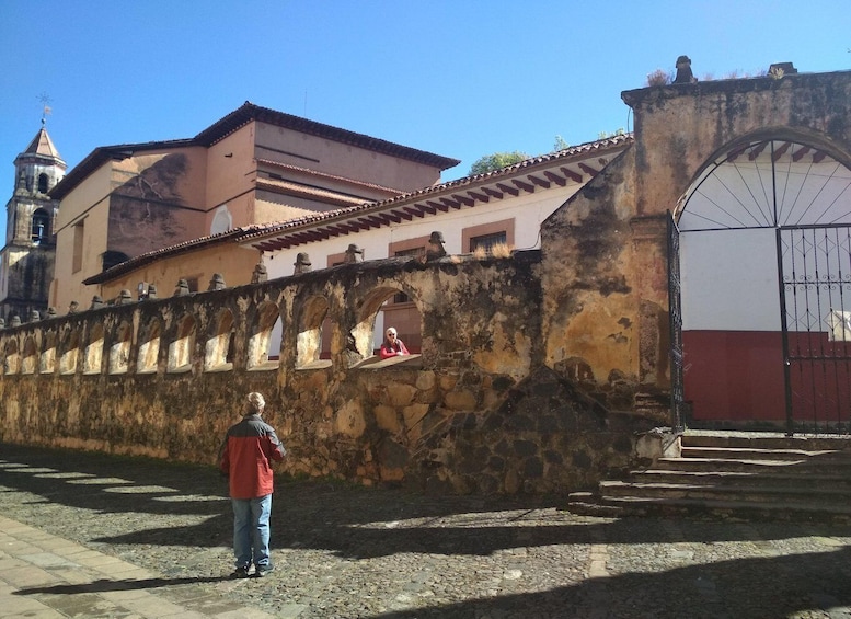 A stone wall with a gate, a person standing in front, and a building with a bell tower in the background. 5