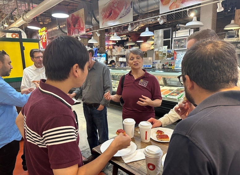 A group of people dining inside a food court, with a woman in a maroon shirt speaking to them. 5