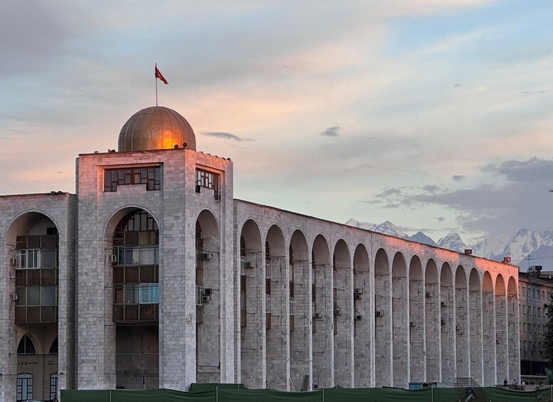 A large, historic building with a dome and arched windows, set against a mountainous backdrop. 1