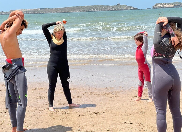 Un grupo de personas con trajes de neopreno en una playa arenosa, con el océano y una isla rocosa distante al fondo. 4