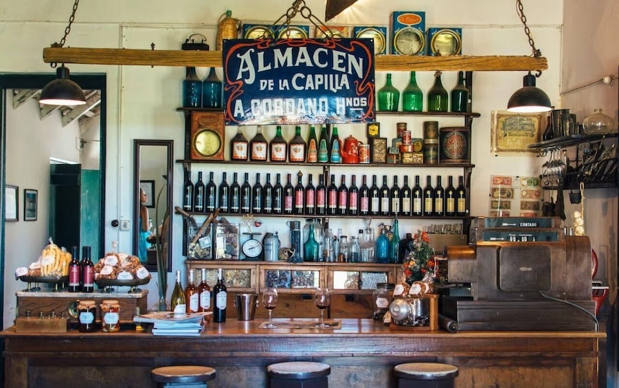 Un bar avec un comptoir en bois rustique, des étagères remplies de bouteilles de toutes sortes et une enseigne indiquant : « Almacen de la Capilla A Cordano Hnos ». 1