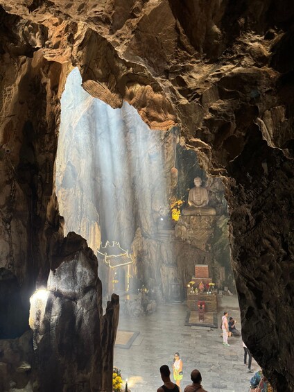 A cave interior with a large rock formation, a Buddha statue, and visitors. 1