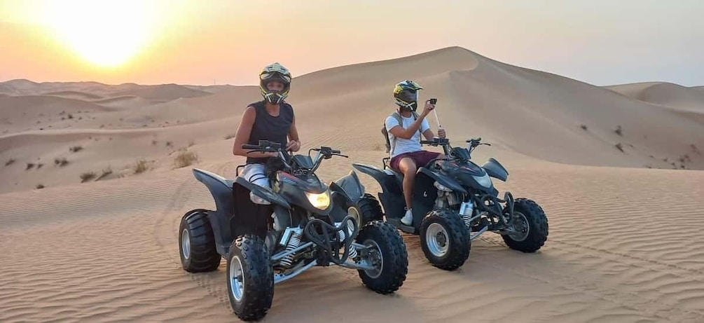 Two people riding ATVs on a desert sand dune during sunset. 1