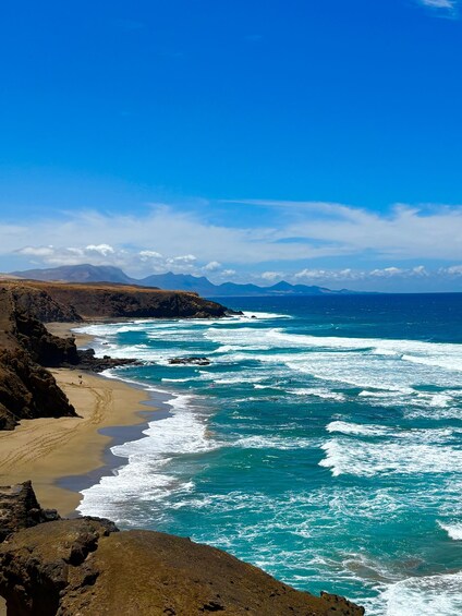 A coastal landscape with rocky cliffs, a sandy beach, and turquoise ocean waves under a clear blue sky. 1