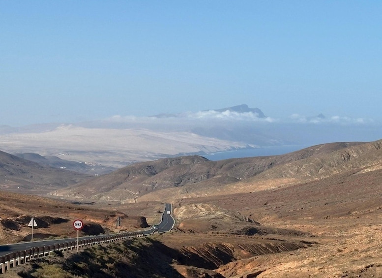 A winding road in a barren landscape with distant mountains and a body of water. 1