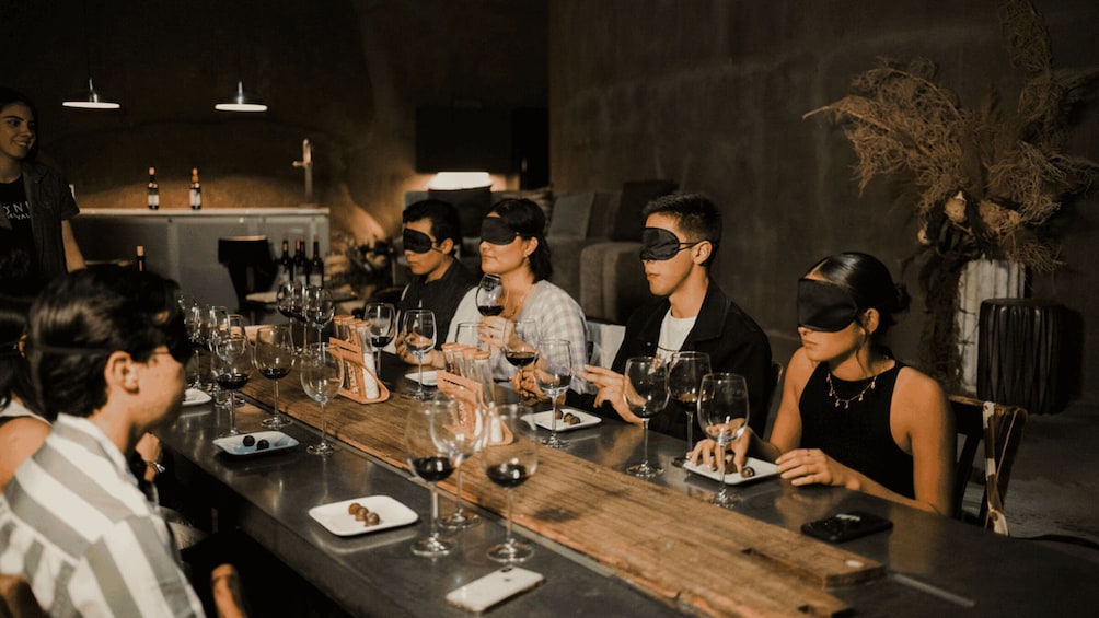 Un groupe de personnes est assis autour d’une longue table en bois, dans une pièce à la lumière tamisée. Devant eux, on peut voir des verres à vin et des assiettes remplies de nourriture. 5