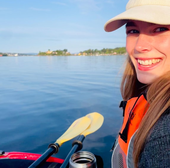 Una persona su un kayak, con un remo, indossa un giubbotto di salvataggio e un cappello, l'acqua è calma e si vede una costa in lontananza. 3
