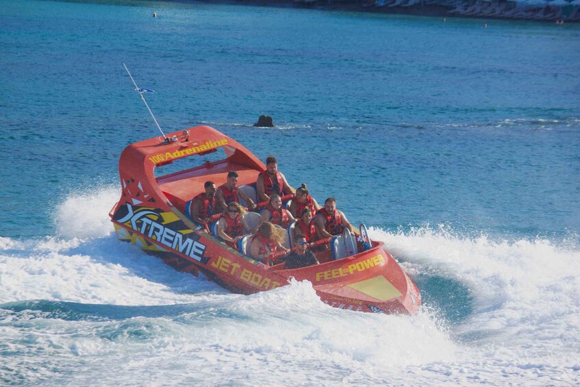 A red jet boat with passengers wearing life jackets, moving through a body of water. 2
