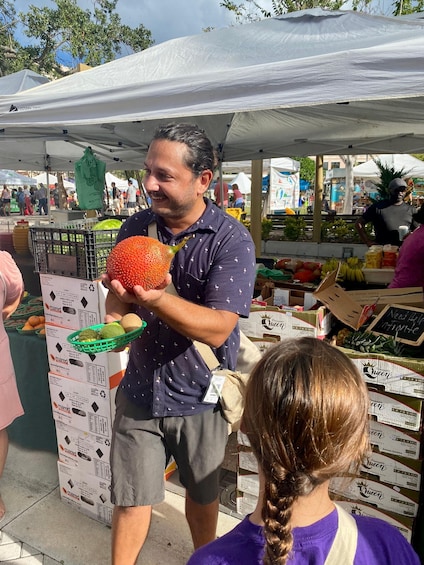 Eine Person hält eine stachelige Frucht an einem Marktstand, wo Kisten mit der Aufschrift 'Queen' und diverse Waren ausliegen. 5