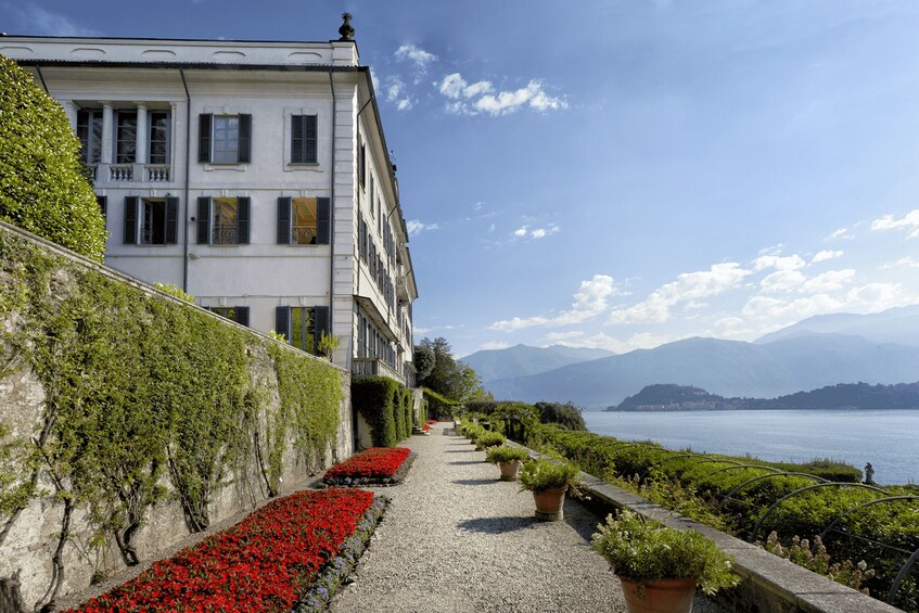 A pathway lined with red flowers and potted plants, leading to a white building with columns and shutters, overlooking a lake with mountains in the background. 2