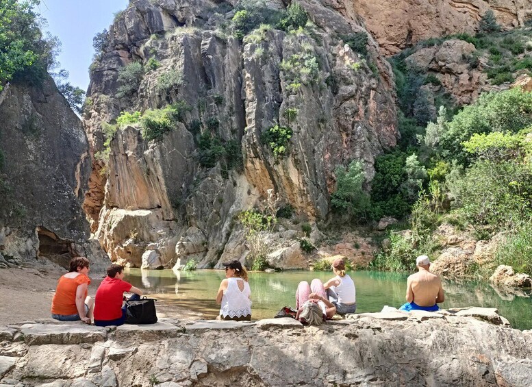 People sitting by a calm river with rocky cliffs in the background. 4