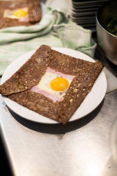 Lautasella on neliönmuotoinen ruoka-annos, mahdollisesti pannukakku tai ohukainen, jonka päällä on paistettu kananmuna ja viipale kinkkua. 5