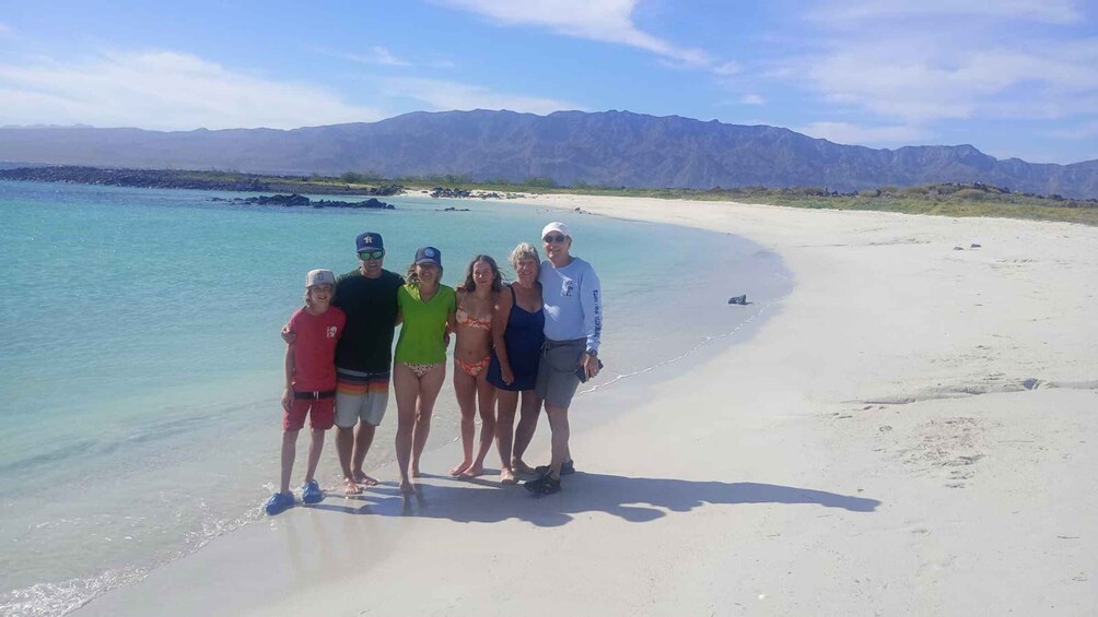 A group of people standing on a sandy beach with clear turquoise water and mountains in the background. 2