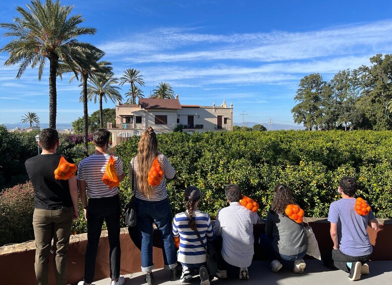 A group of people standing and sitting outdoors, some holding orange fruits, with palm trees and a house in the background. 2