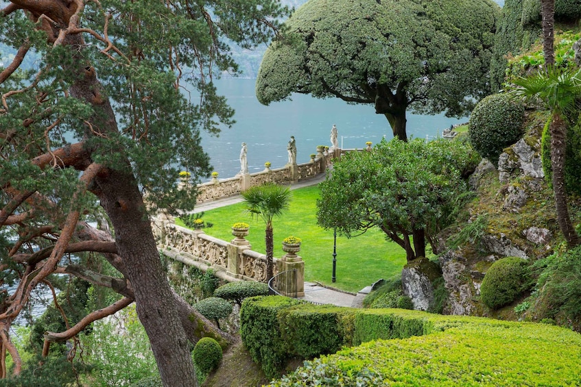 A landscaped garden with a stone pathway, statues, and a large tree overlooking a body of water. 1