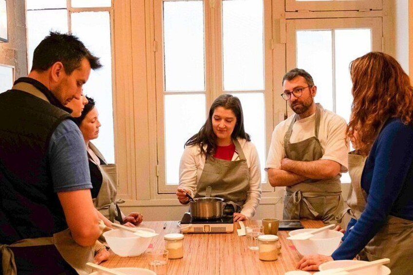 A group of people in a kitchen, some wearing aprons, with a woman cooking at the center. 2