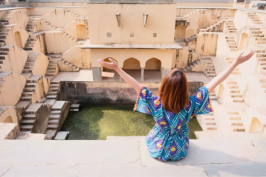 A person sitting on a ledge with arms outstretched, overlooking a traditional water body with steps leading down to it. 2