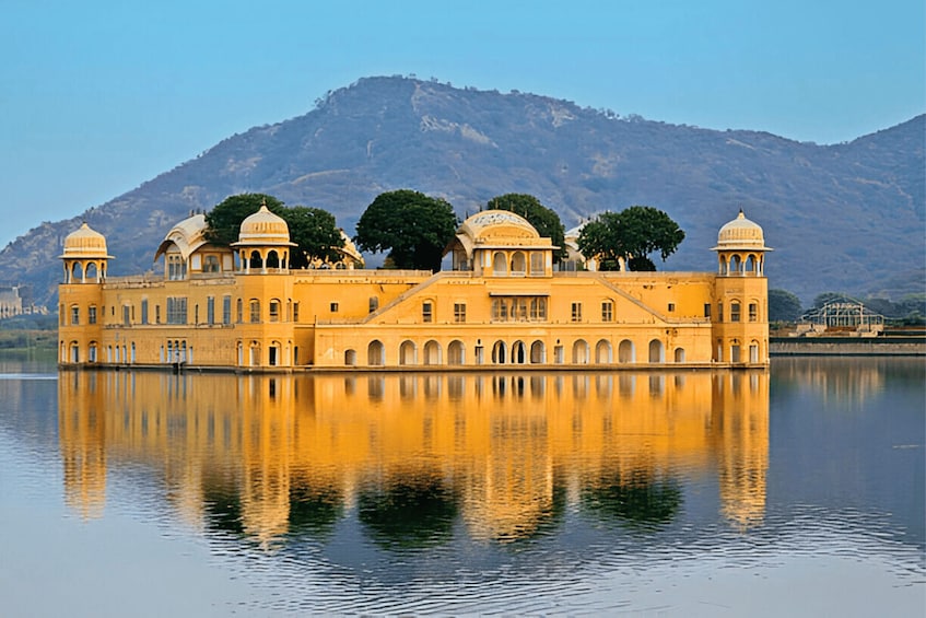 A large, yellow building with multiple domes and arched windows, reflecting on calm water, with mountains in the background. 4