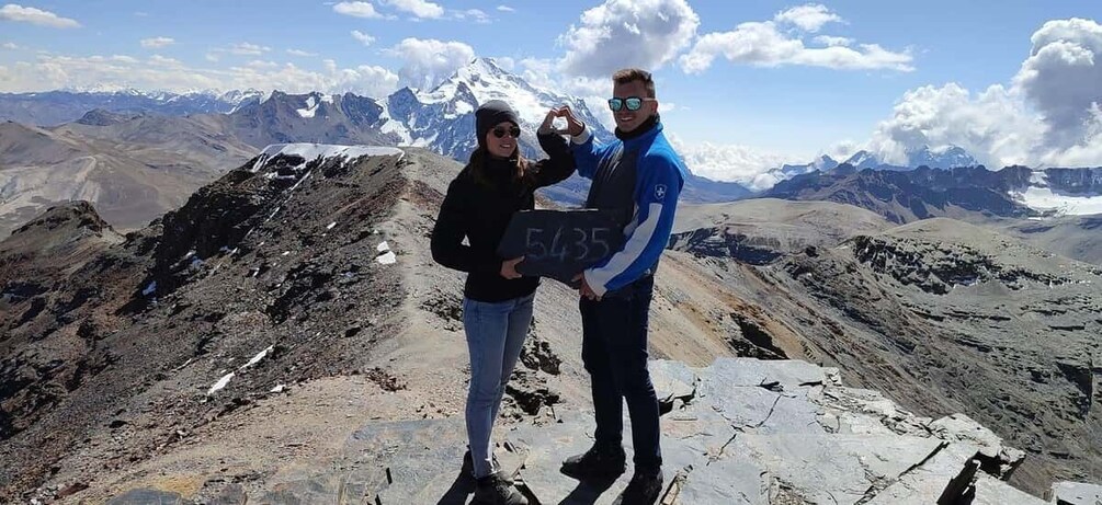 Two people standing on a mountain peak with a sign reading '5435' and a view of snow-capped mountains in the background. 1