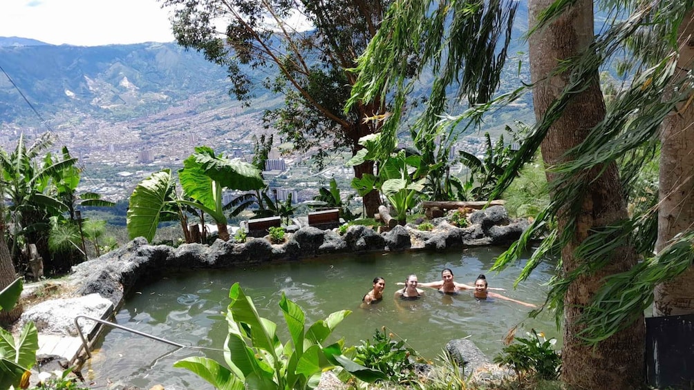 A natural hot spring with people swimming, surrounded by lush vegetation and a cityscape in the background. 1