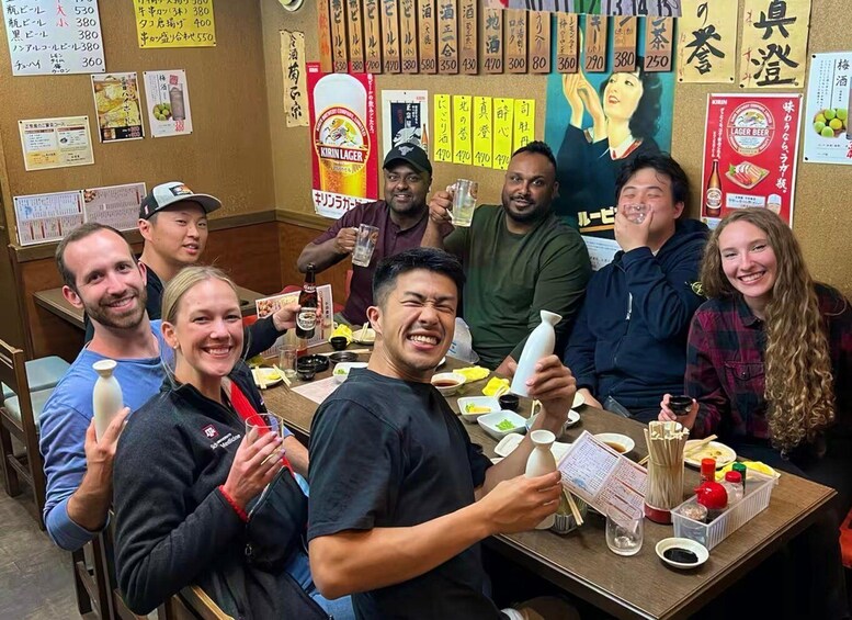 A group of people sitting around a table with food and drinks, in a room with various posters and signs on the walls. 1