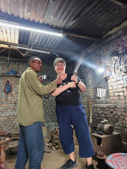 Two people in a rustic room with brick walls, a metal roof, and various tools and objects. 3