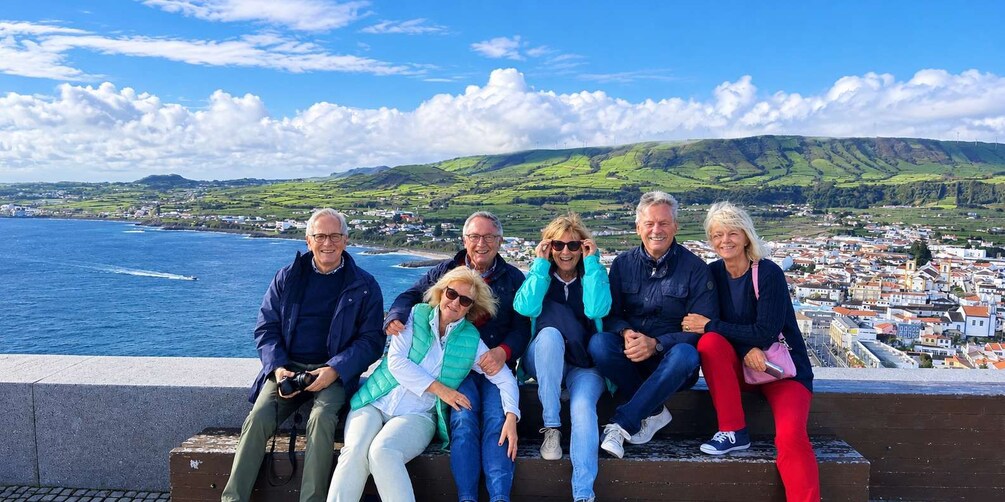 A group of people sitting on a ledge overlooking a coastal town with green hills in the background. 4