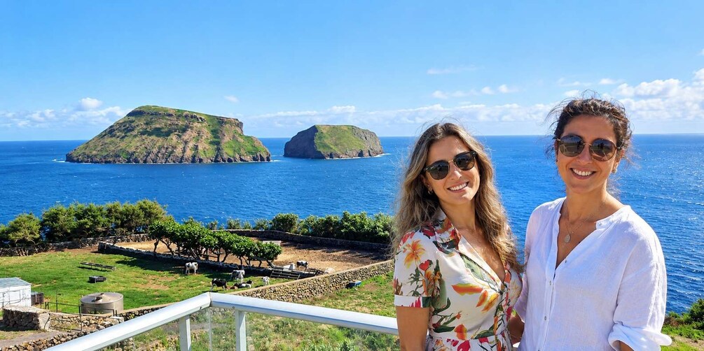 Two people standing on a balcony overlooking a coastal landscape with green hills and a clear blue sea. 1