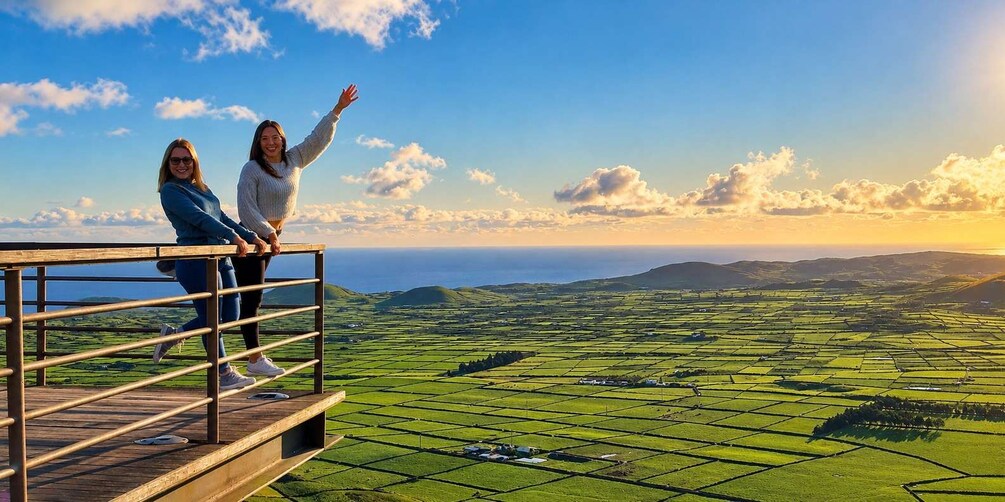 Two people standing on a wooden platform overlooking a green landscape with fields and distant hills. 2