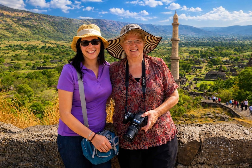 Two people standing by a stone wall with a camera, overlooking a mountainous landscape with a mosque in the distance. 1