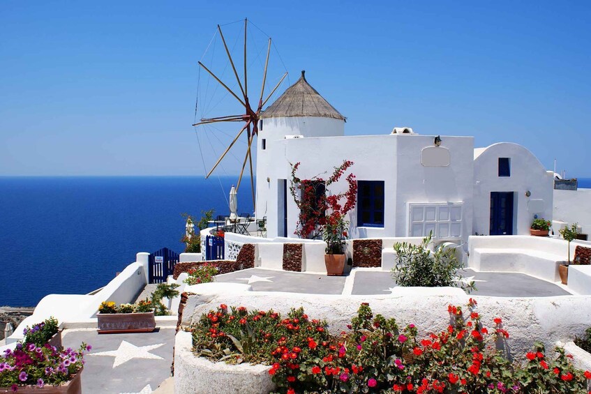 Un edificio blanco con un molino, flores rojas y el mar al fondo. 5