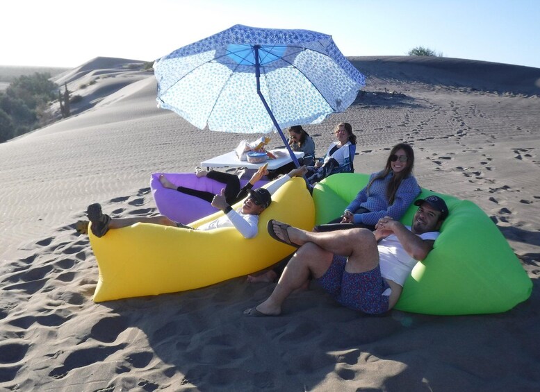 People relaxing on colorful inflatable loungers on a sandy beach with a blue umbrella providing shade. 5