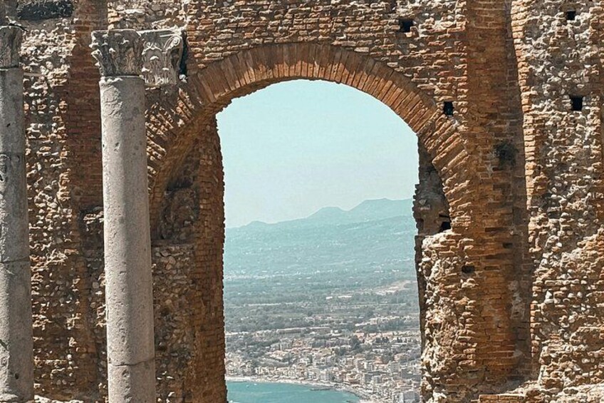 An ancient stone archway with columns, overlooking a coastal cityscape with mountains in the distance. 5