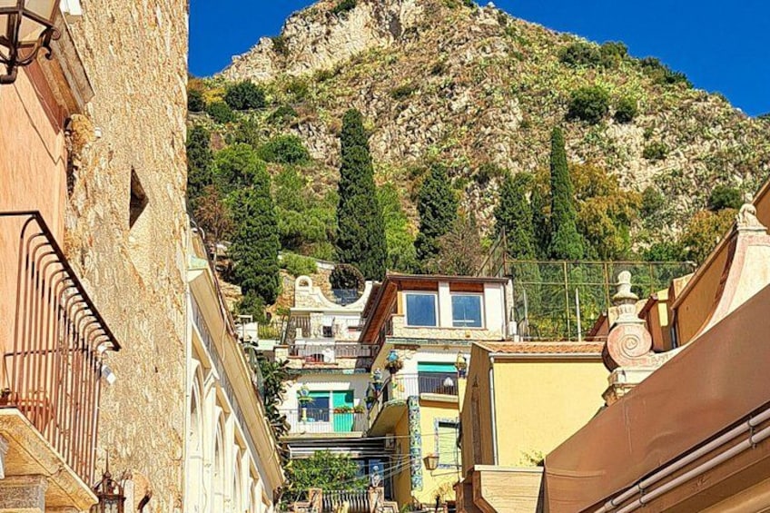A narrow street with buildings, a mountain in the background, and a clear blue sky. 2