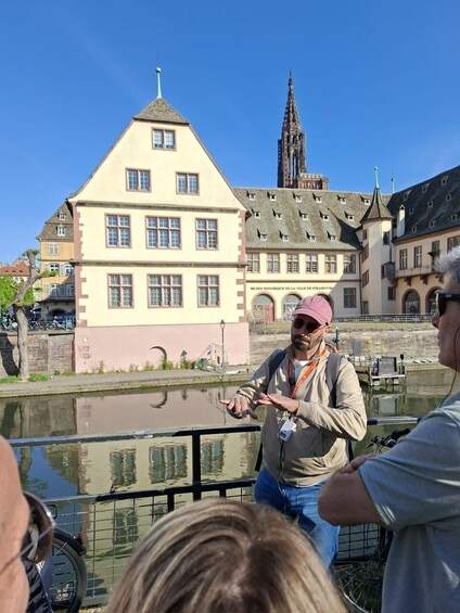 A person gesturing in front of a historic building with a tower, a canal, and a bicycle in the foreground. 2