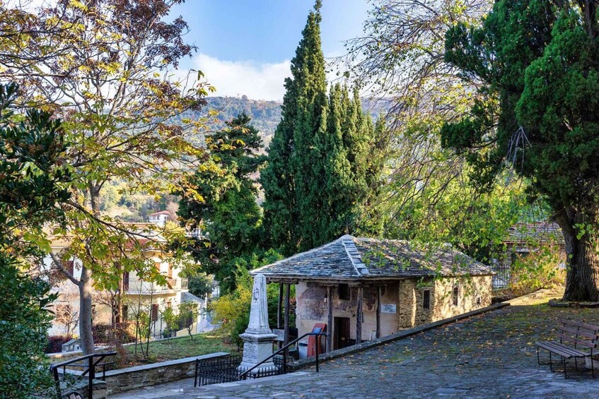 A stone pathway leading to a small stone building with a tiled roof, surrounded by trees and a distant village. 4