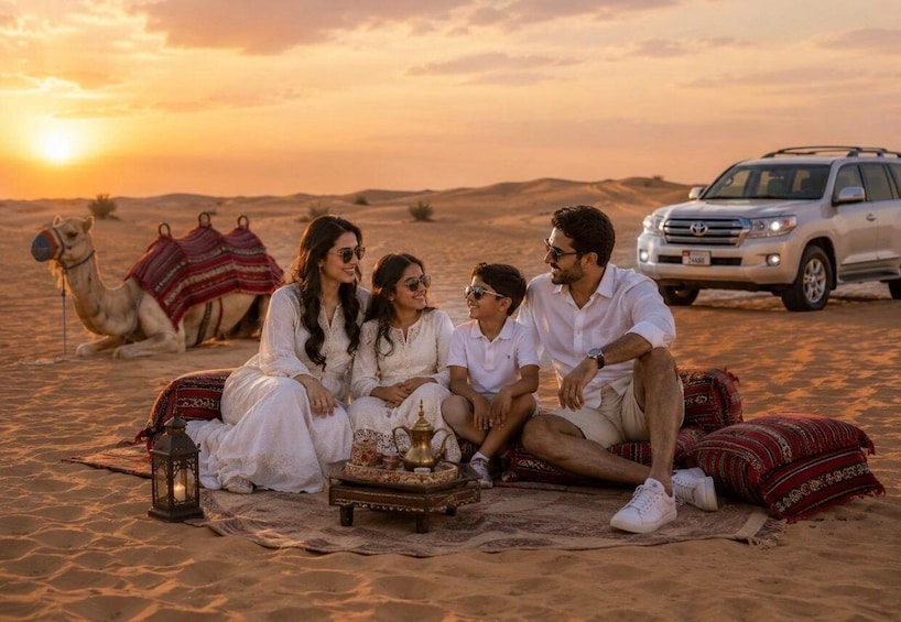 A group of people sitting on a blanket in a desert with a camel and a car in the background. 2