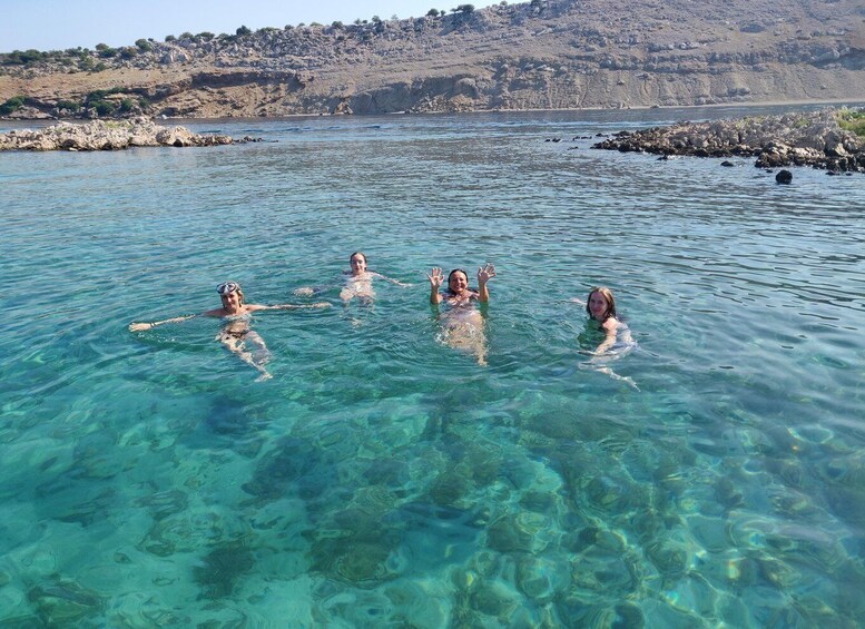 Four people swimming in clear blue water with rocky terrain and hills in the background. 1