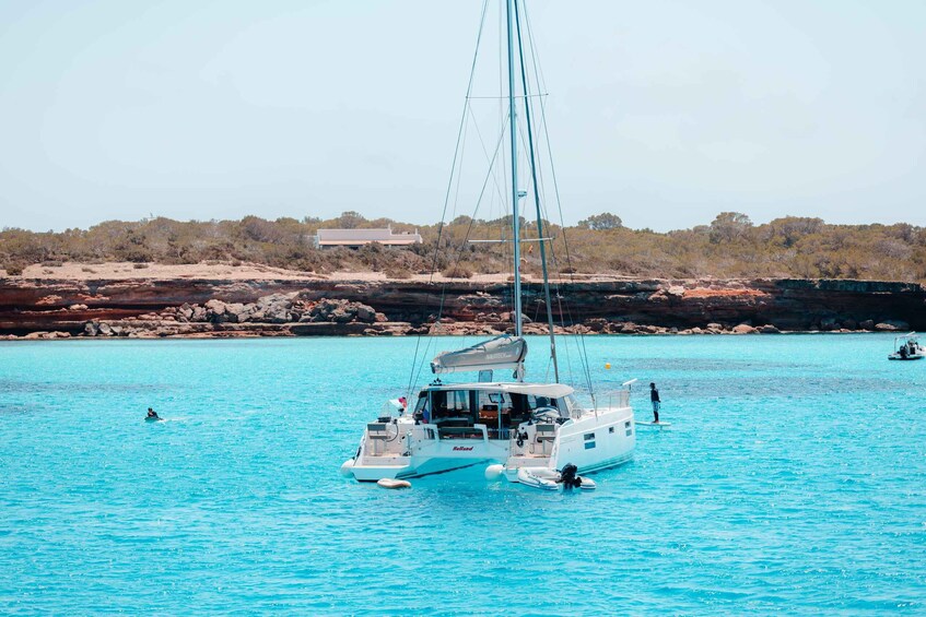 A white sailboat with a cabin and a person standing on the deck, anchored in clear turquoise water with a rocky shoreline and sparse vegetation in the background. 5