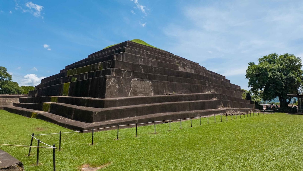 Eine große, stufenförmige Pyramidenstruktur mit flacher Spitze, umgeben von einer Rasenfläche und einem Zaun. 3