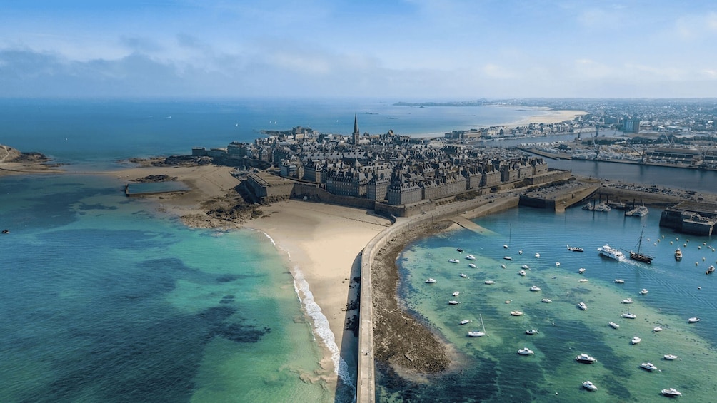Vista aérea de una ciudad costera con un castillo destacado, una playa de arena y un puerto deportivo con numerosos barcos. 1