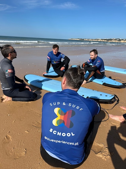 Tre personer sidder på en strand med surfbræder, iført blå våddragter med 'SUP & SURF SCHOOL' på ryggen. 1