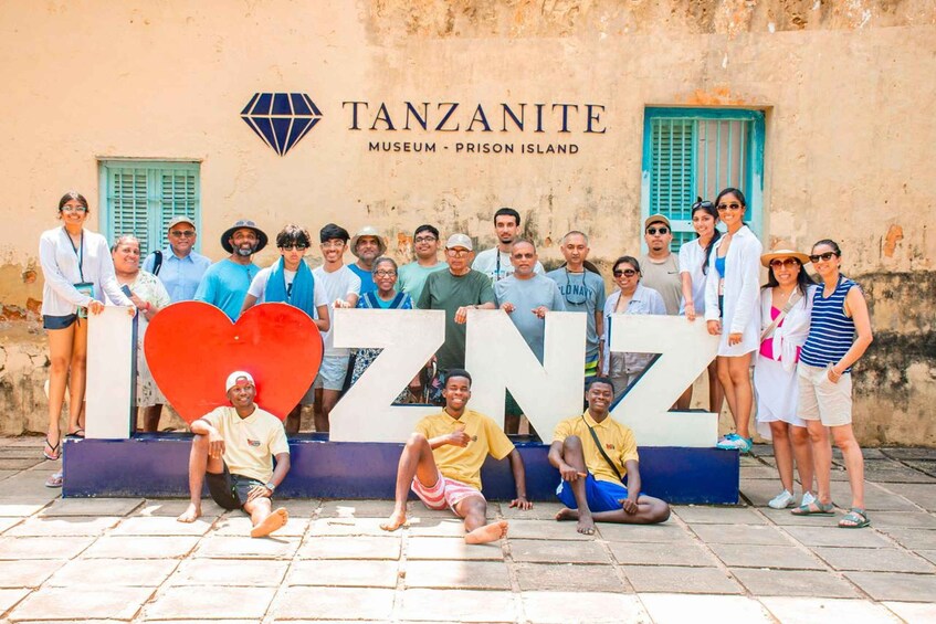 A group of people posing in front of a sign that reads 'TANZANITE MUSEUM - PRISON ISLAND' with a large heart-shaped sign that says 'I ❤️ NZ'. 1