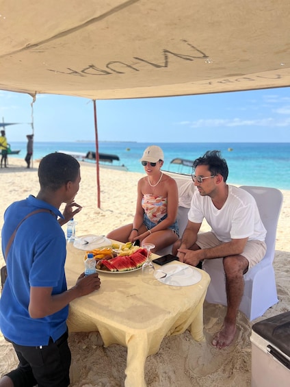 A beach scene with a table set for a meal, people sitting around it, and the ocean in the background. 4