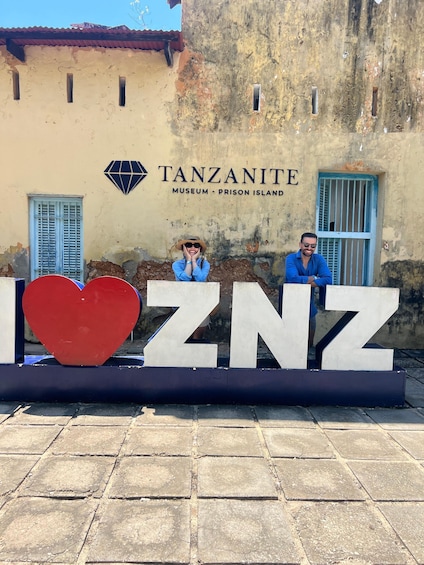 A sign with the words 'TANZANITE' and 'MUSEUM - PRISON ISLAND' on a weathered wall, with two people standing behind a large heart-shaped sign. 2