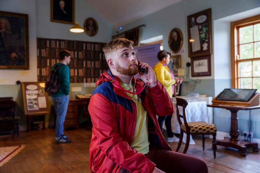 A person in a red jacket sitting in a room with wooden floors, bookshelves, and a table with a computer. 1