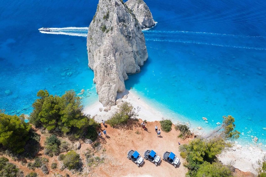 A coastal landscape with a prominent rock formation, clear blue waters, and a sandy beach with parked vehicles. 1